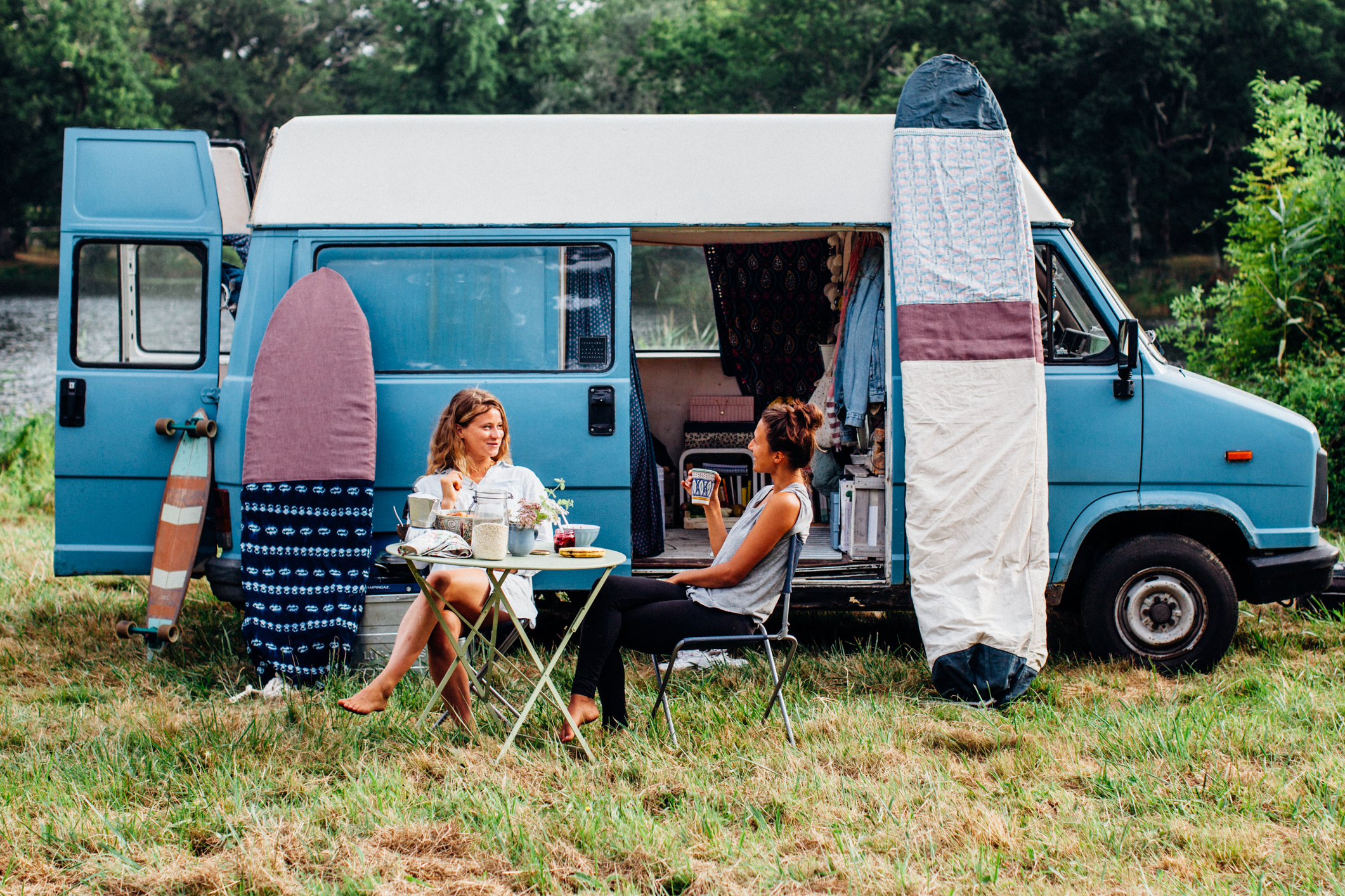 Deux femmes autour d'une table face à un van et deux planches de surf entourées d'arbre